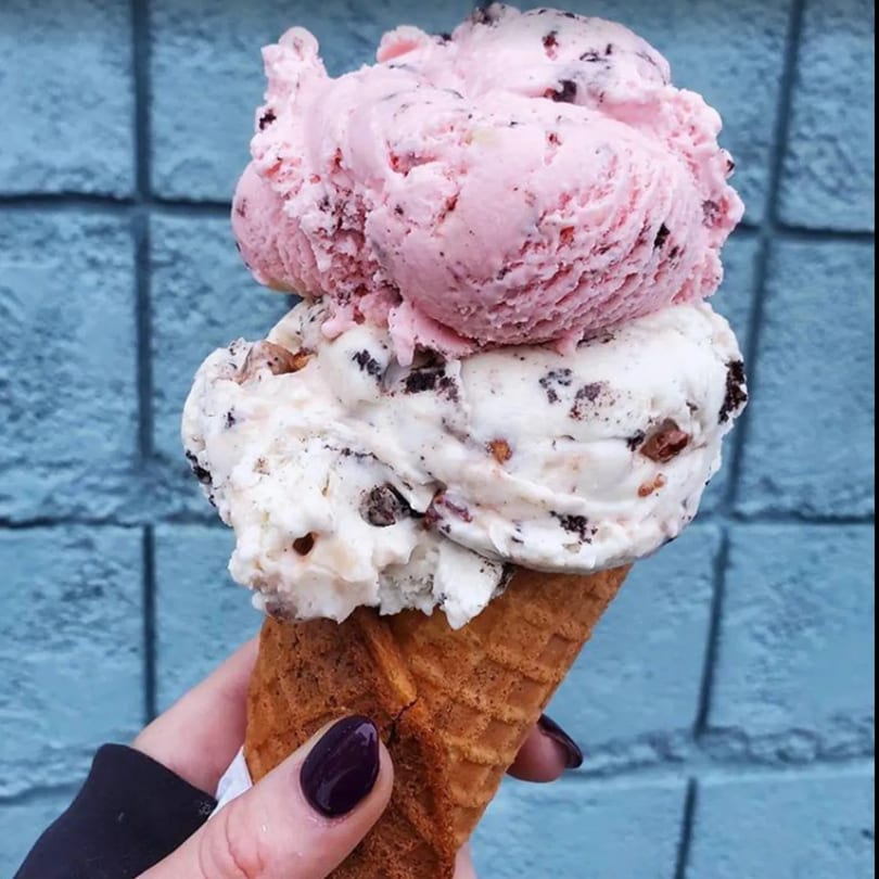 A hand holds a Grand Ole Creamery waffle cone with scoops of pink and cookies and cream ice cream.