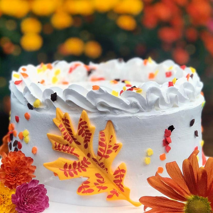 White cake with autumn leaf decorations, colorful sprinkles, and fresh flowers.