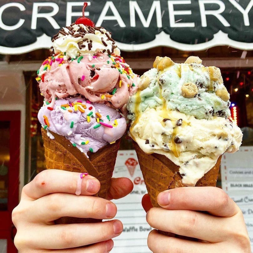 Two hands holding waffle cones with scoops of Grand Ole Creamery ice cream, topped with sprinkles and syrup.
